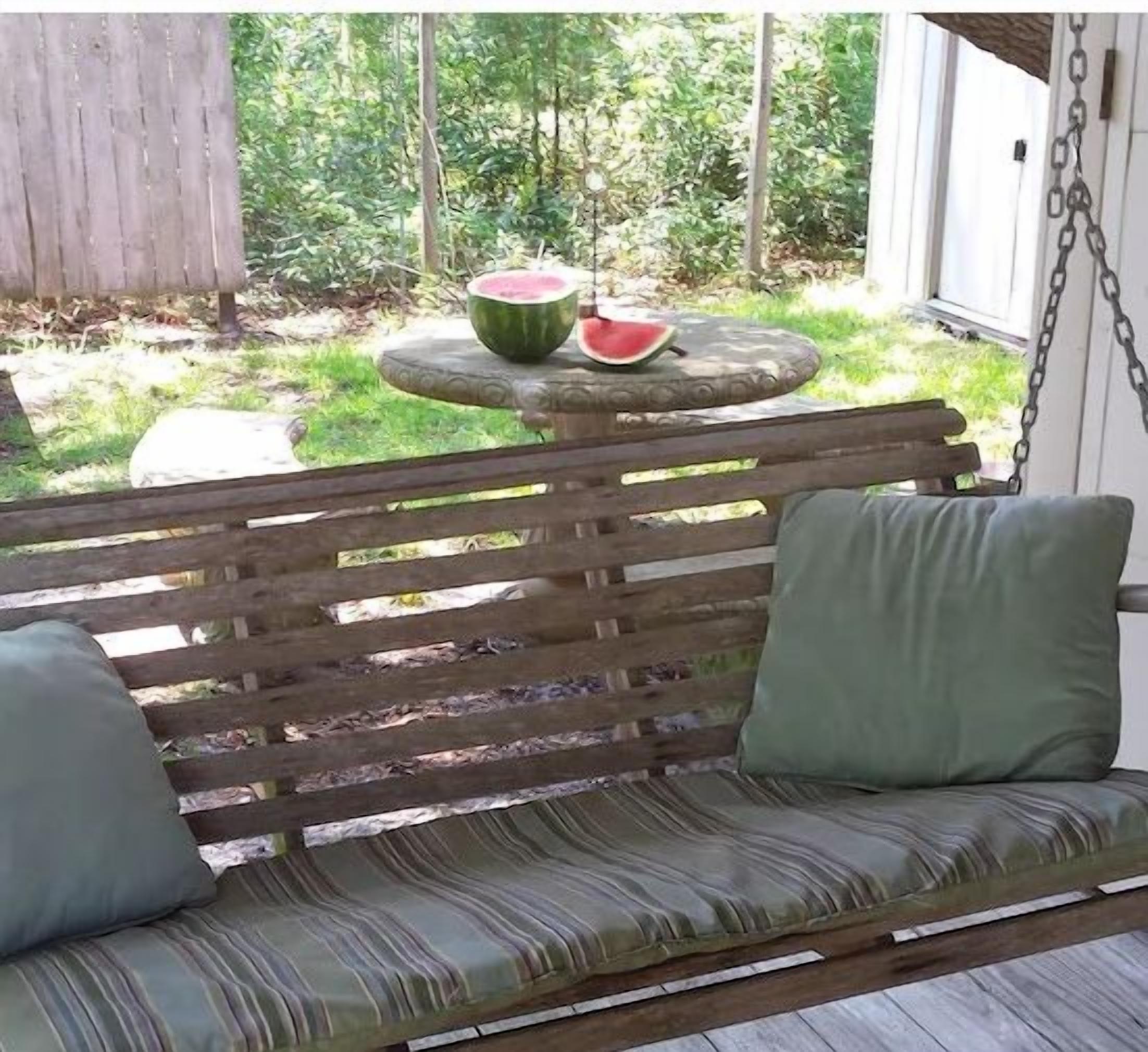 Eine Holzbank mit Kissen, ein kleiner Tisch mit Wassermelone und ein Maschendrahtzaun in einem Hinterhof.
