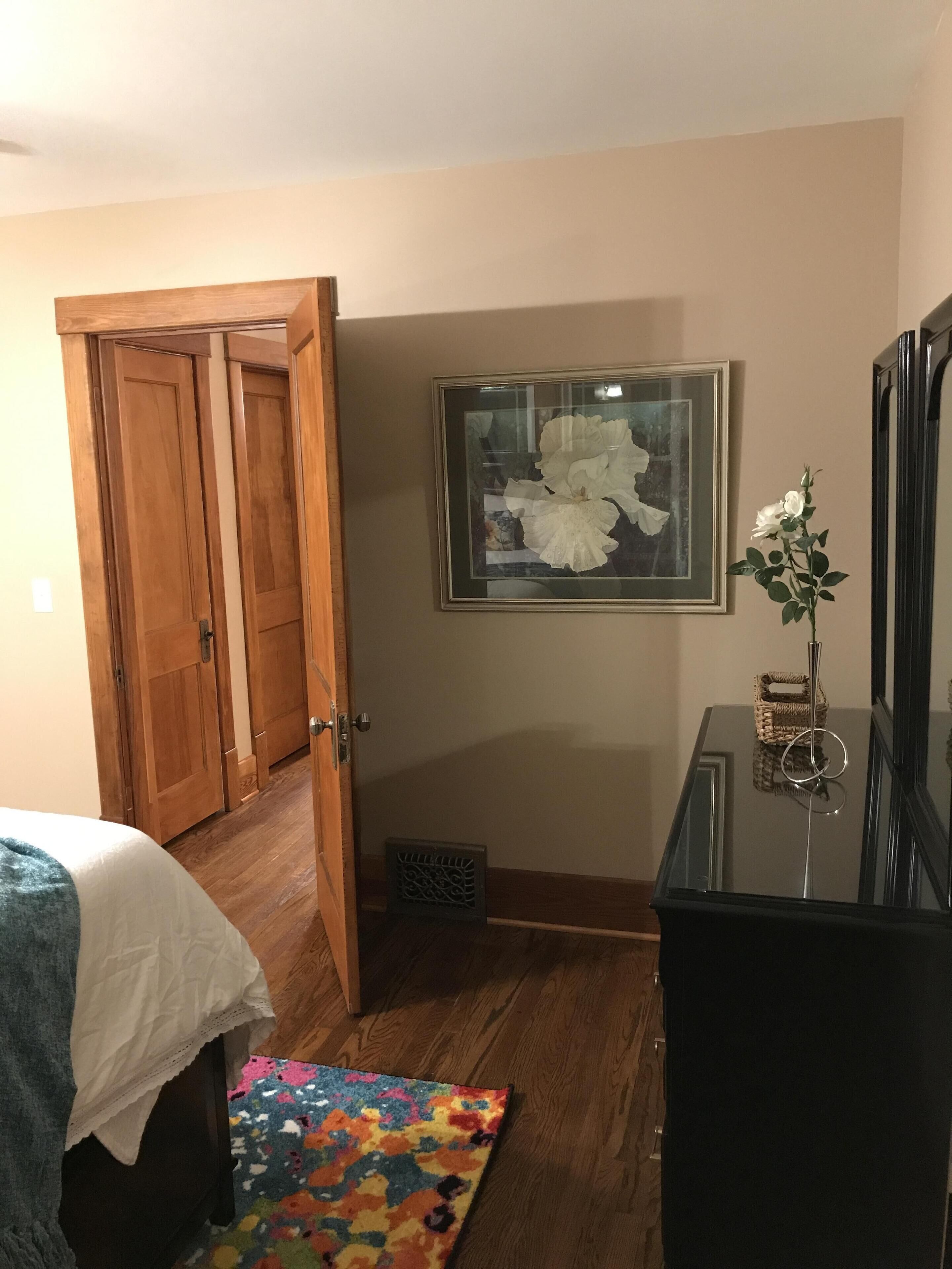 Quarto com cama, tapete colorido, guarda-roupa de madeira, quadro emoldurado, cômoda e espelho.