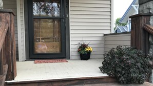 Una escalera de madera que conduce a una puerta principal con marco negro, una pequeña planta en maceta y un arbusto grande en el porche.