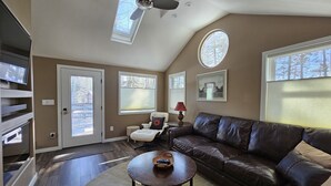 Un salon spacieux avec un canapé en cuir, une table basse ronde et un ventilateur de plafond. De grandes fenêtres laissent entrer la lumière naturelle et offrent une vue sur l’extérieur.