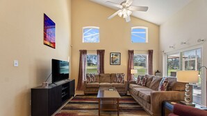 A living room with a sofa, a coffee table, a TV, and a ceiling fan. - Golf Course View Villa! Just 25 mins to Disney Magic (Davenport)