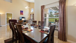 A dining area with a glass table, wooden chairs, and a view of a tennis court through large windows. - Golf Course View Villa! Just 25 mins to Disney Magic (Davenport)