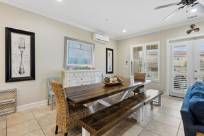 A dining area with a wooden table, wicker chairs, and a bench. There is a white cabinet with drawers and a framed picture above it.