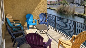 En balkon med loftventilator, flere farverige Adirondack-stole, et lille bord og udsigt til vand og bygninger i baggrunden.