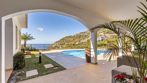 A pool area with a view of the sea, a garden, and a hillside with houses. - Luxurious house with sea and mountain views (Arco da calheta)