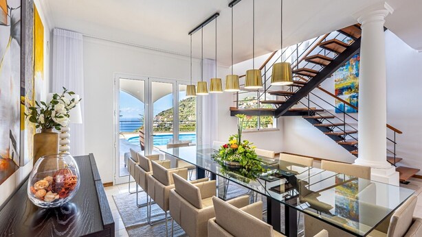 A modern dining area with a glass table, beige chairs, and a view of the sea. - Luxurious house with sea and mountain views (Arco da calheta)