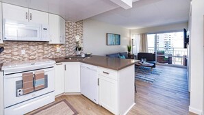 Een moderne keuken met witte kasten, een magnetron en een betegelde spatwand. Er is een zithoek met een blauwe bank en uitzicht op de stad door grote ramen.