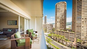 Een balkon met rieten meubilair, een bank en een klein tafeltje, met uitzicht op een stadsbeeld met hoge gebouwen en een weg.