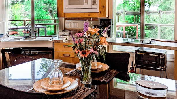 A kitchen with a microwave, a dining table with chairs, a vase with flowers, and large windows with a view of greenery. - Visit award winning wineries in our valley in tricked out Yurt on the NF River. (Hotchkiss)