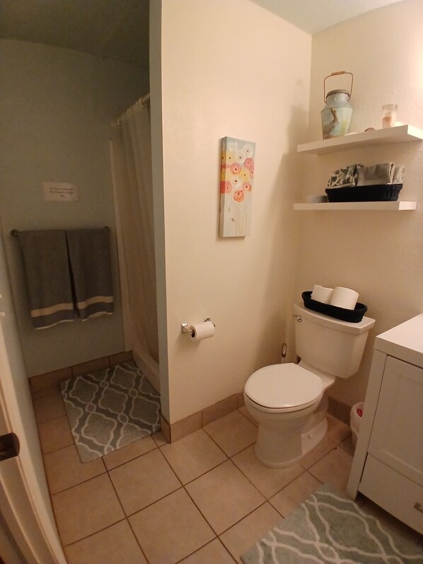 A bathroom with a toilet, a shower curtain, a patterned rug, a toilet paper holder, and a shelf with a basket.