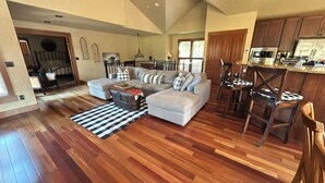 Amplia sala de estar con un sofá seccional en forma de L, una mesa de centro y un tapete a rayas. Se aprecia un comedor con sillas de madera y una cocina al fondo.