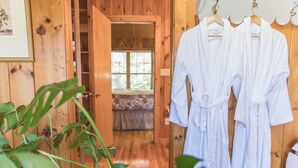 A wooden interior with a wooden door leading to a room, white robes hanging on a rack, and a plant in the foreground. - Rustic Romantic Bayberry Cottage at Autumn Ridge (Lexington)