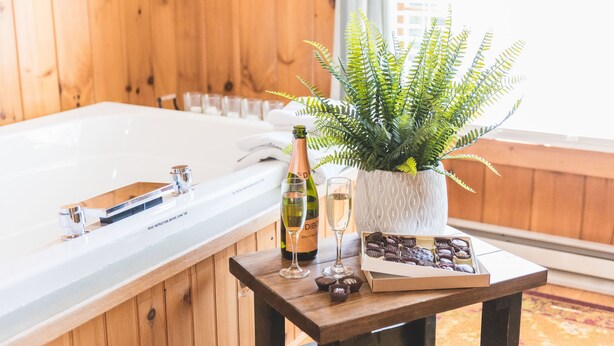 Interior de una cabaña de madera con una bañera grande, una botella de champán, dos copas y una caja de bombones sobre una mesita.