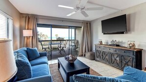 Sala de estar com sofá, mesa de centro e TV fixada na parede. Varanda com vista para o oceano.