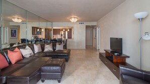A modern living room with a sectional sofa, a flat-screen TV, and a dining area in the background. - On The Beach !!!  2 Minutes to Exquisite Dinning and Shopping (Hallendale Beach)
