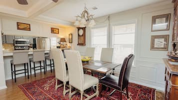 A dining area with a wooden table, four chairs, a chandelier, and a kitchen in the background.