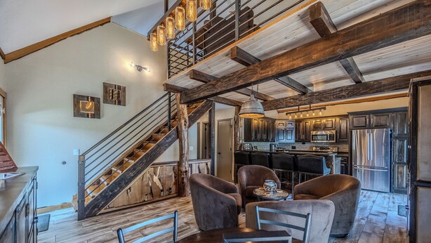 Une cuisine spacieuse équipée d’appareils modernes, un coin repas avec une table et des chaises en bois, et un escalier rustique menant à l’étage supérieur.