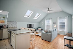 A modern kitchen with a central island, a dining area with a table and chairs, a grey sofa, and a ceiling fan.