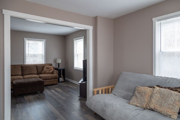 A living room with a brown sectional sofa, a grey sofa, and a wooden bench. There is a television on a stand and a lamp on a side table.