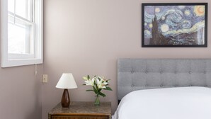 Een slaapkamer met een bed, een nachtkastje met een lamp en bloemen, een schilderij aan de muur, en een raam.