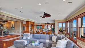 A spacious living room with a gray sectional sofa, ottomans, and a coffee table. There is a dining area with wooden furniture and a kitchen in the background. - Last minute SPECIAL’s @ Bella Vita moorage, chef's kitchen, optional add 2 bdr ! (Seattle)