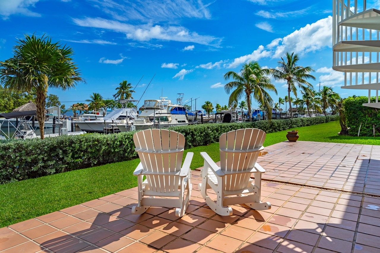 Two Adirondack chairs on a tiled patio with a view of a marina and palm trees.