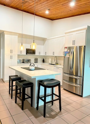 A modern kitchen with white cabinets, a stainless steel refrigerator, a built-in oven, and a central island with stools.