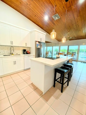 Een moderne keuken met witte kasten, een groot eiland met barstoelen en een uitzicht op de buiten omgeving door grote ramen.