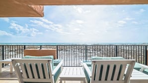 Un balcon avec des chaises blanches et une table, donnant sur une plage sous un ciel dégagé.