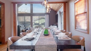 A dining area with a long table set for a meal, large windows, and a view of the outside.