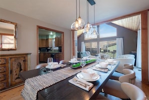 Un espace repas avec une longue table dressée pour le repas, une cuisine en arrière-plan et une vue sur l’extérieur grâce à de grandes fenêtres.