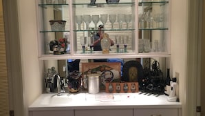 Una cocina con armarios blancos, una encimera con varios utensilios y un expositor de vidrio sobre los armarios que contiene cristalería y objetos decorativos.