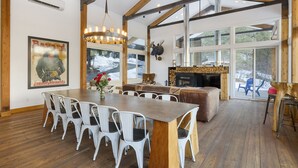 A spacious dining area with a long wooden table and white chairs, a chandelier, and a large window with a view of a snowy landscape. - 5-Star Lodge \ Groups Welcome  \ 1 Mile from Downtown (Leavenworth)