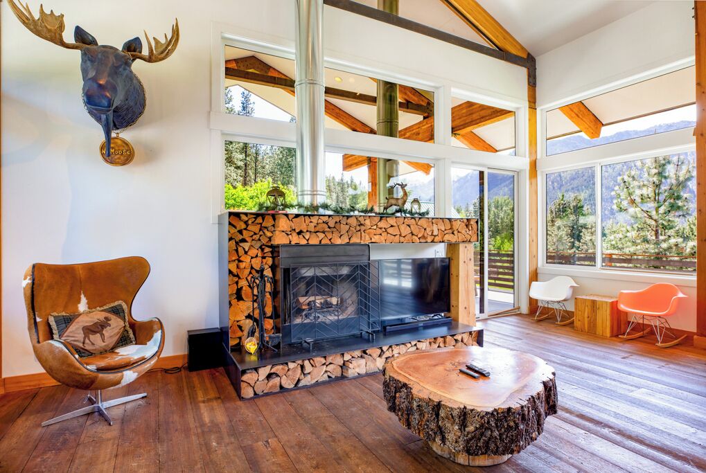 Un salón acogedor con chimenea de piedra, vigas de madera, una cabeza de alce montada en la pared y un ventanal con vistas a la vegetación.