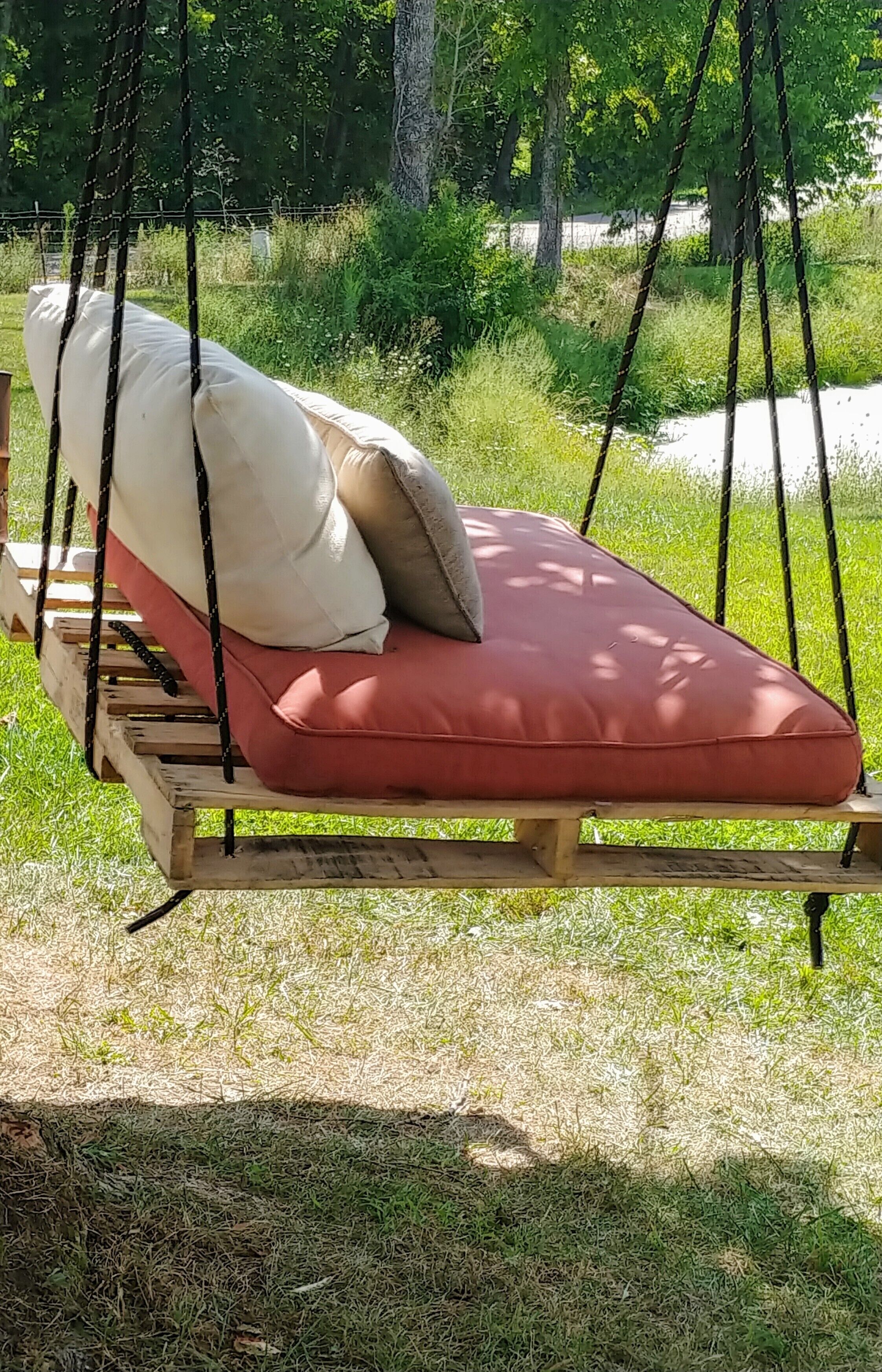 Un fauteuil suspendu en bois, doté d’un coussin rouge et de coussins beiges, installé dans un espace extérieur verdoyant, avec des arbres en arrière-plan.