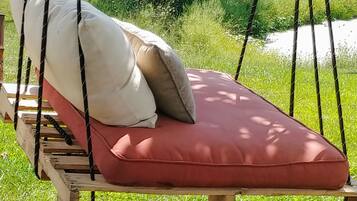 Un fauteuil suspendu en bois, doté d’un coussin rouge et de coussins beiges, installé dans un espace extérieur verdoyant, avec des arbres en arrière-plan.