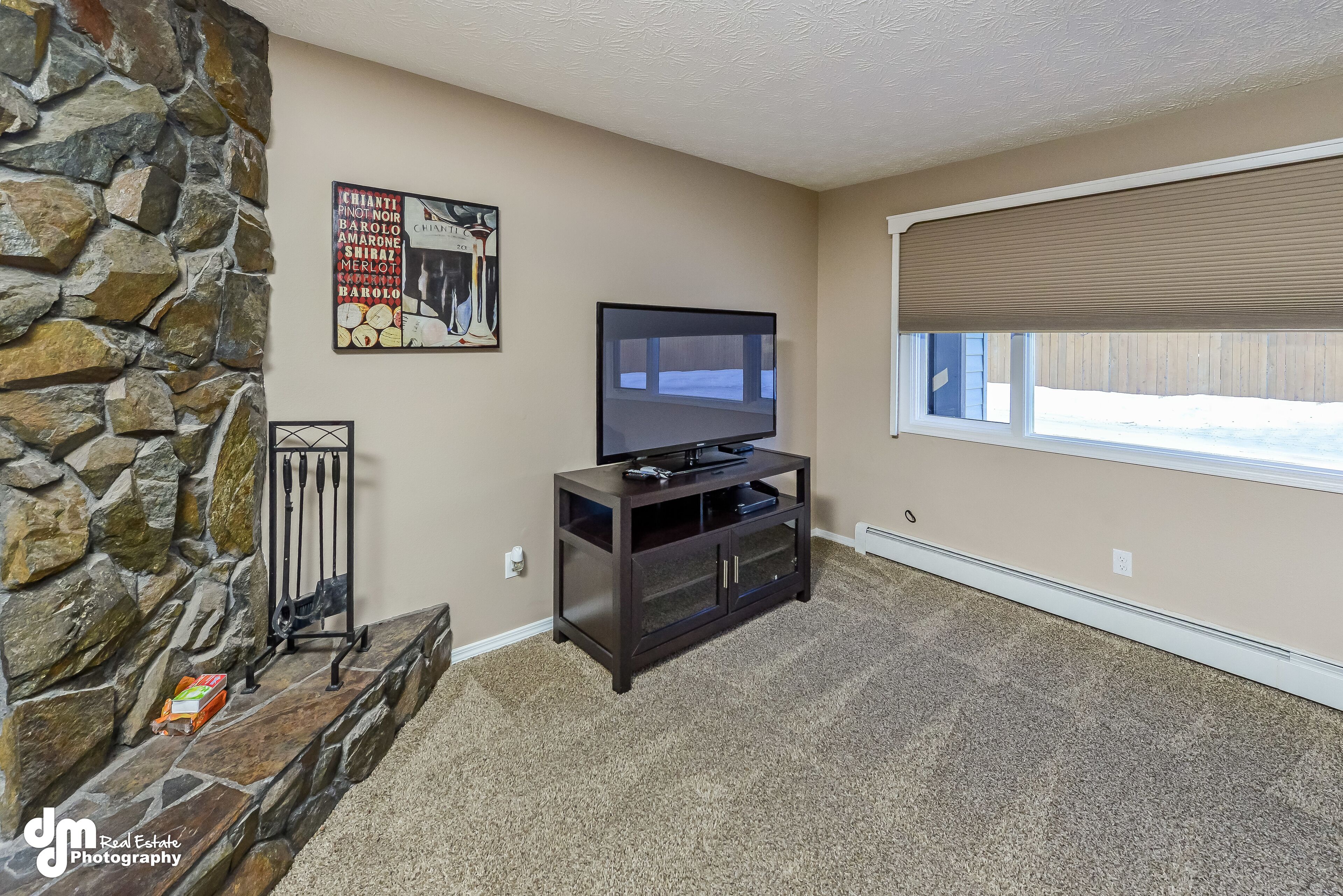 A living room with a stone wall, a flat-screen TV on a dark wooden stand, a window with a beige blind, and a small table with a box.