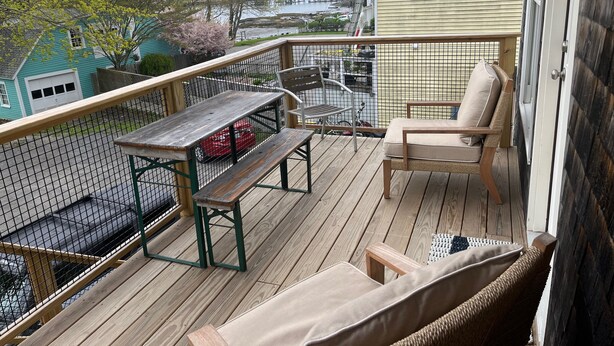 Een houten terras met een tafel en stoelen, uitzicht op een waterpartij, en een huis op de achtergrond.