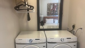Een wasruimte met twee Samsung wasmachines, een witte ventilator en een spiegel die een kamer met een bed en raam weerspiegelt.