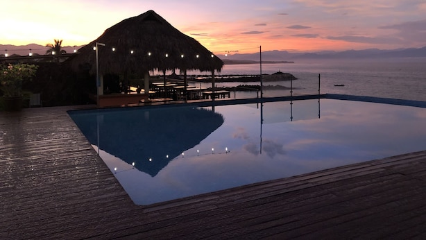 Área da piscina com estrutura de palha, deck de madeira e vista para o mar ao pôr do sol.