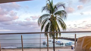 Uma varanda com vista para a praia, uma palmeira e barcos na água.