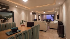 Uma sala de estar moderna com área de jantar, apresentando uma janela grande com vista para o mar, um vaso de planta e uma televisão de tela plana na parede.