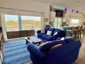 A living room with a large window, a blue sofa, a coffee table, and a dining area with wicker chairs. - #1 Reviewed Home on Navarre Beach Forever Views 33' Pool Outdoor Bar/TVs Hot Tub (Navarre)
