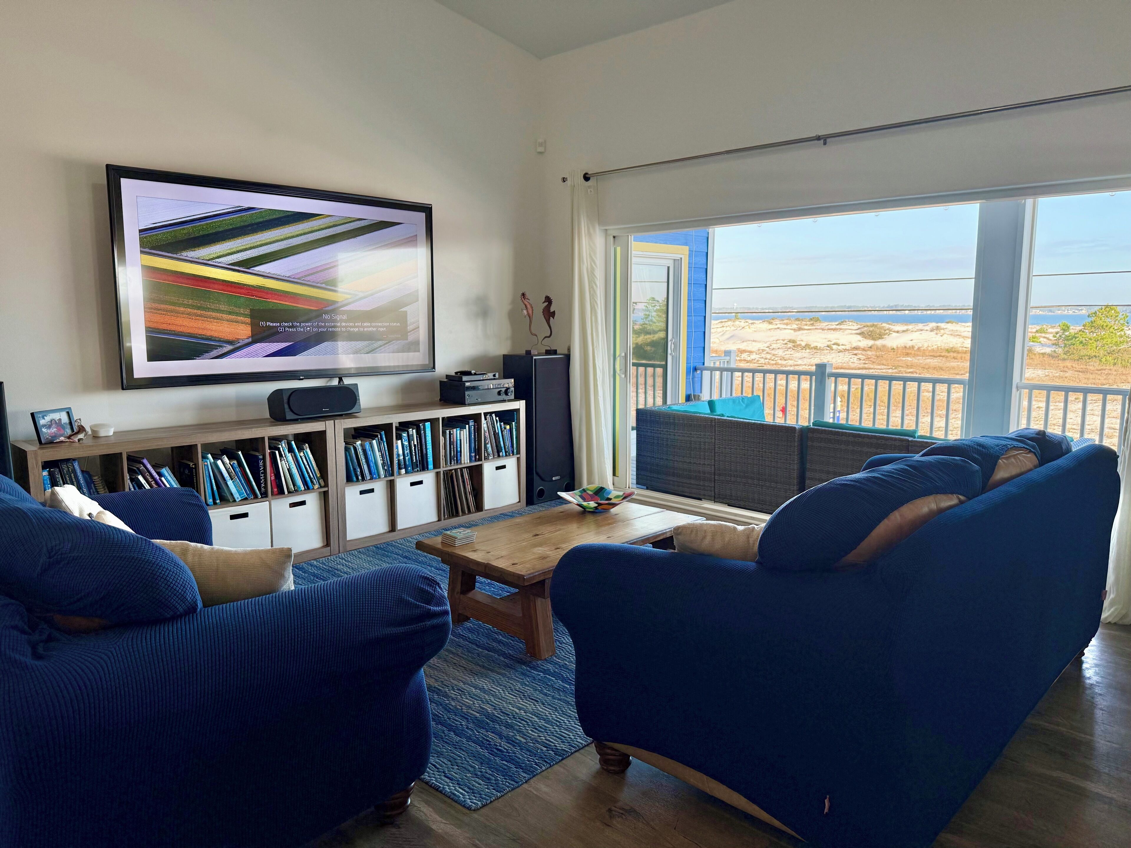 A living room with a large TV, bookshelves, a coffee table, and a view of the beach.