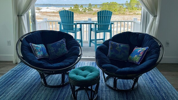 Una habitación con dos sillas azul oscuro y un otoman a juego, una mesita y vistas a la playa y al océano a través de un ventanal grande.