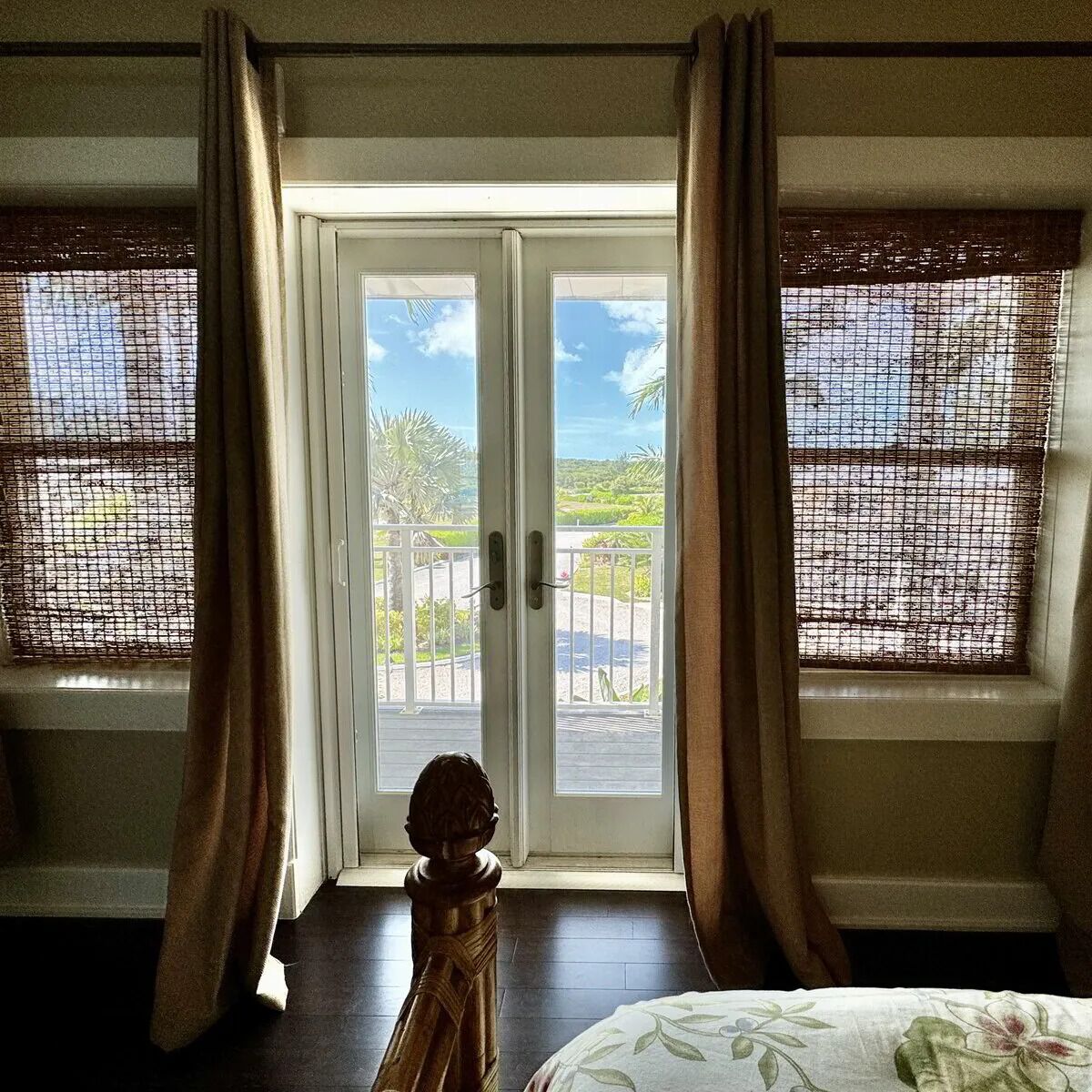 A room with a bed, a wooden chair, and a view through a glass door to an outdoor area with a railing and greenery.