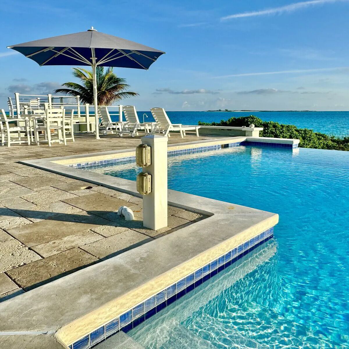 A pool area with lounge chairs and an umbrella, surrounded by a tiled deck and palm trees.