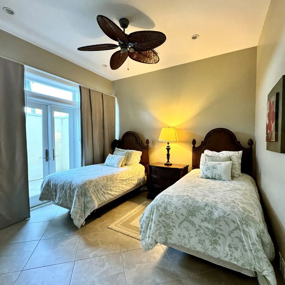 A hotel room with two beds, a ceiling fan, a nightstand with a lamp, and a view through a sliding glass door.