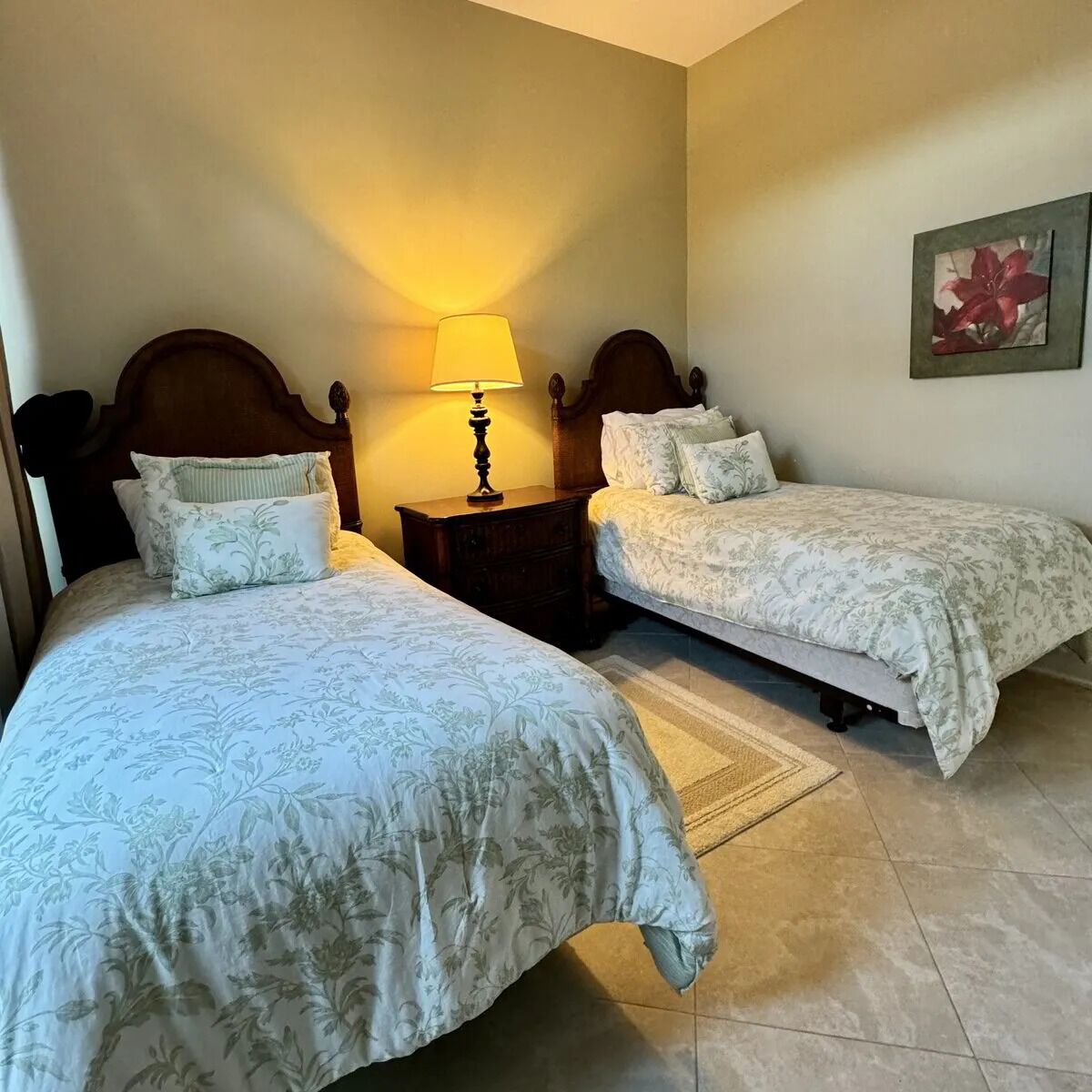 Une chambre avec deux lits, une table de chevet avec une lampe et un tableau représentant des fleurs accroché au mur.