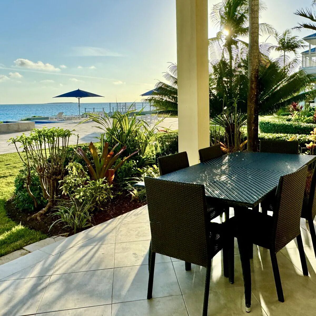 Une terrasse avec une table et des chaises, entourée de plantes tropicales et offrant une vue sur l’océan.
